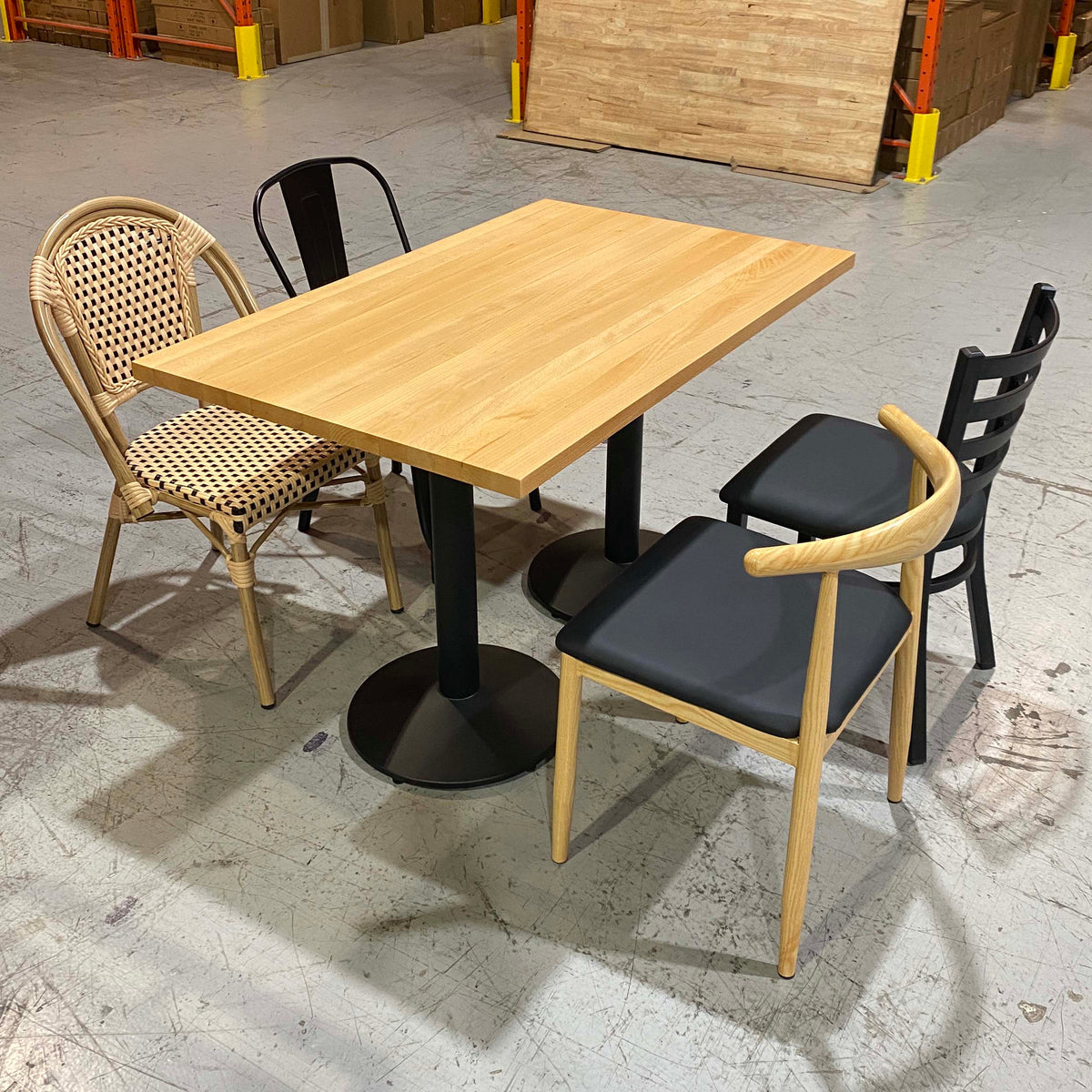 A square wooden dining table with black pedestal legs, surrounded by four chairs in a warehouse showroom setting. Three chairs have light natural wood frames with black cushions, and one has a woven rattan back and seat.