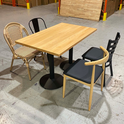 A square wooden dining table with black pedestal legs, surrounded by four chairs in a warehouse showroom setting. Three chairs have light natural wood frames with black cushions, and one has a woven rattan back and seat.