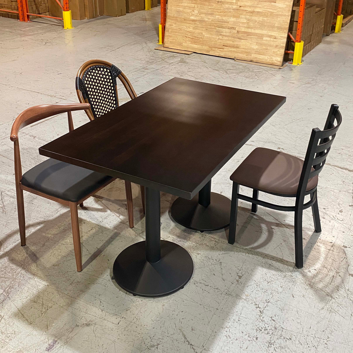 A dark brown rectangular dining table with a black pedestal base, accompanied by two chairs in a warehouse showroom setting.