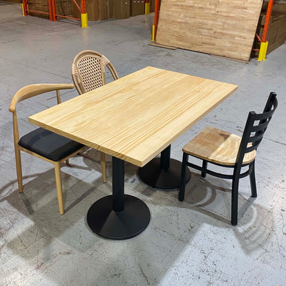 A light natural wood dining table with a black pedestal base, shown with two chairs in a warehouse showroom setting.