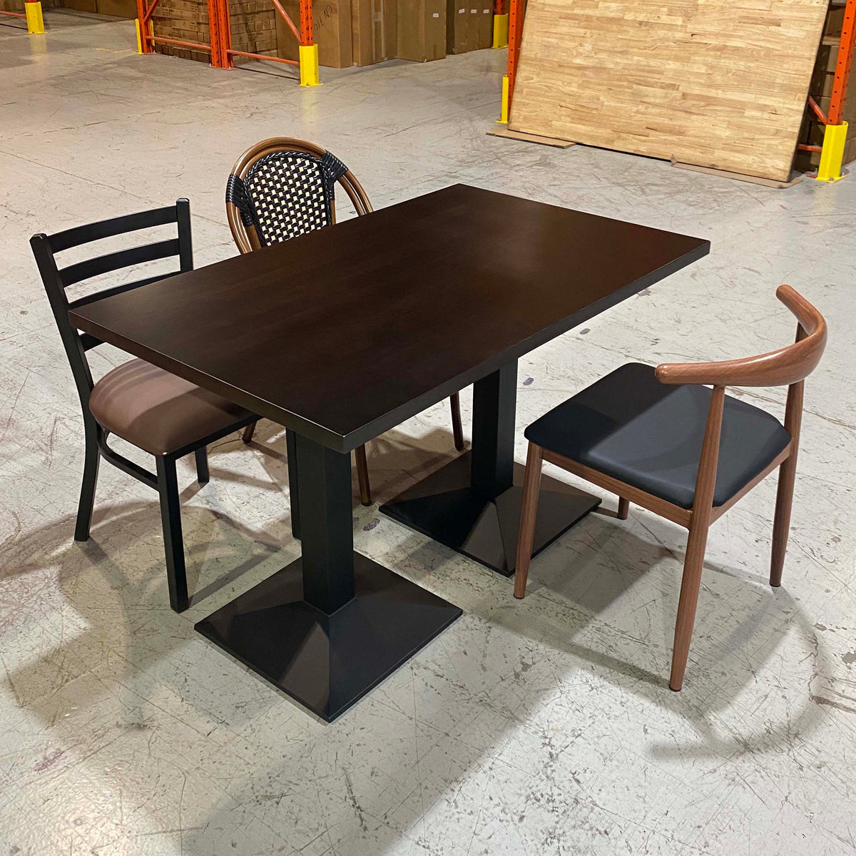 A dark brown rectangular restaurant table with black pedestal legs, shown with three chairs in a warehouse showroom setting.