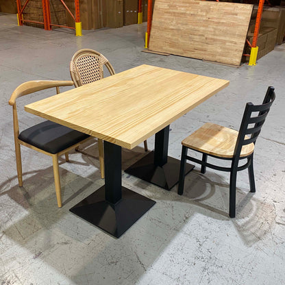 A rectangular wooden dining table with black pedestal bases, shown with two chairs in a warehouse-like showroom. One chair has a light wood frame and black seat, the other has a woven back, and a third black chair is placed beside the table.