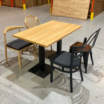 A rectangular wooden dining table with black metal legs is shown with three chairs in a warehouse-like setting. Two chairs have black seats and backs, and one has a woven cane back.