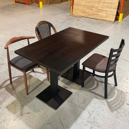 A dark brown rectangular restaurant table with a square pedestal base, shown with two chairs in a warehouse showroom setting.