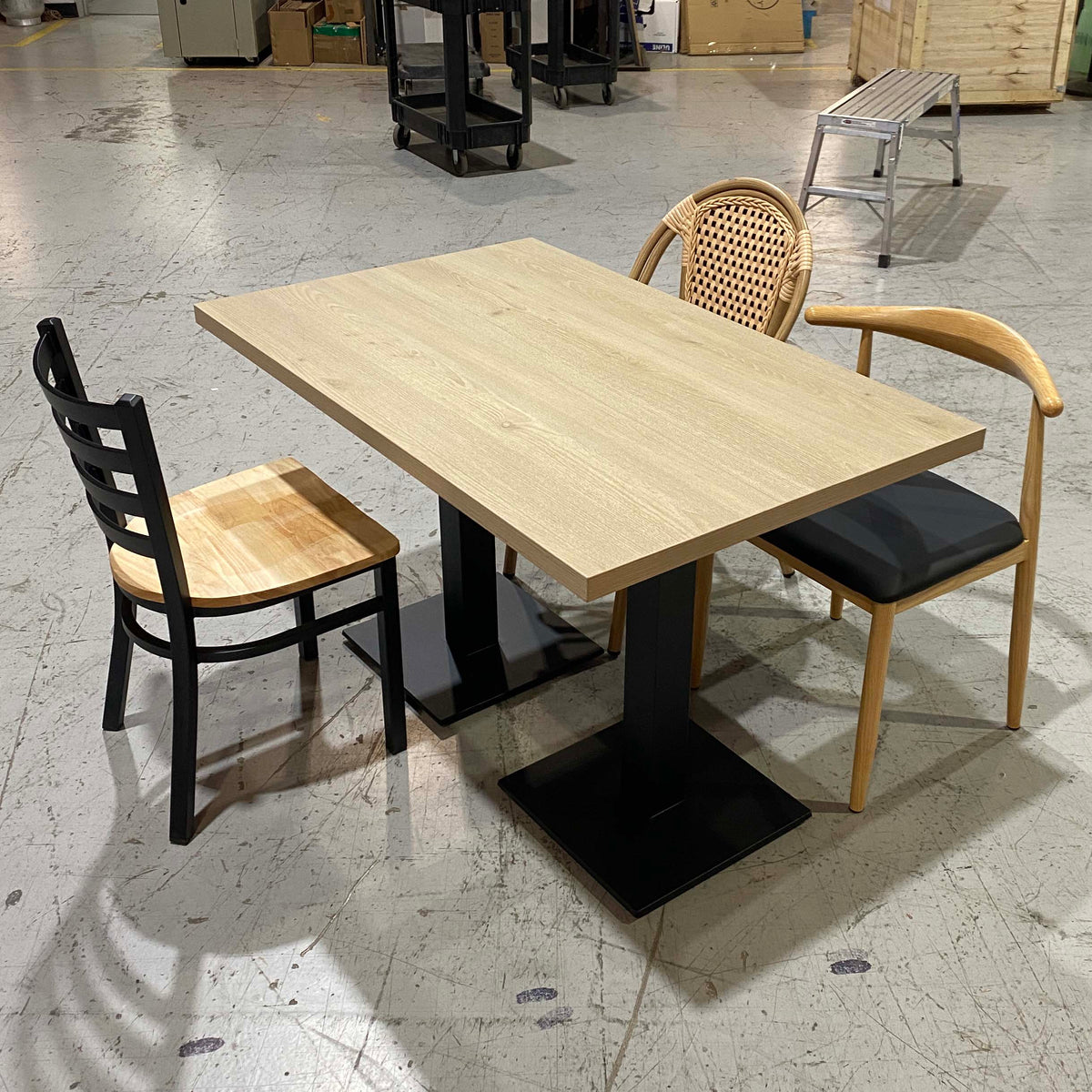 A rectangular light wood dining table with black pedestal bases, shown with two chairs in a warehouse-like setting. One chair has a black frame and light wood seat, and the other has a woven back, light wood frame, and black cushion.