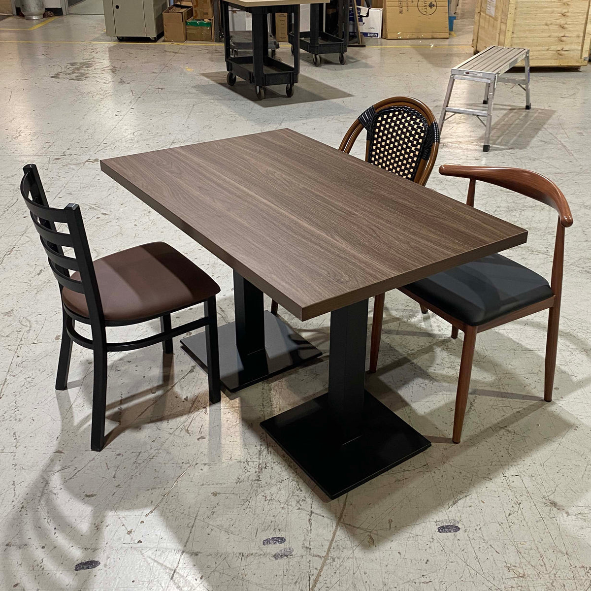 A rectangular restaurant-style table with a dark woodgrain top and black pedestal base, surrounded by two chairs in a warehouse or showroom setting.