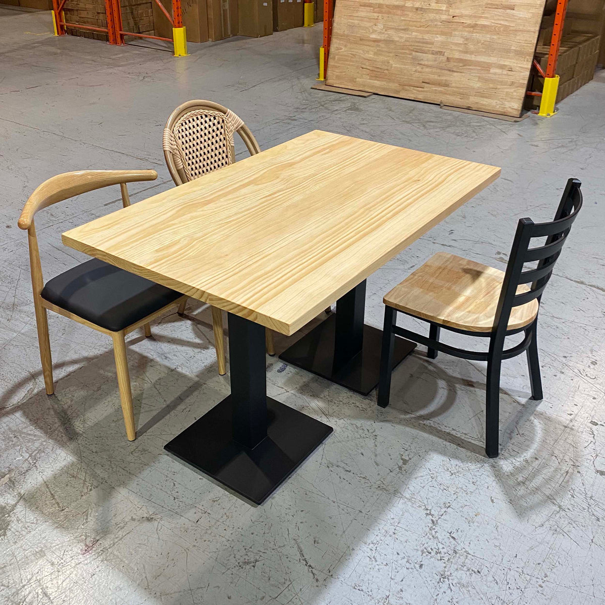 A rectangular light wood dining table with a black pedestal base, shown with two chairs in a warehouse showroom. One chair has a light wood frame and black seat, and the other has a woven cane back and light wood seat.