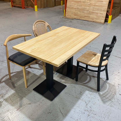 A rectangular light wood dining table with a black pedestal base, shown with two chairs in a warehouse showroom. One chair has a light wood frame and black seat, and the other has a woven cane back and light wood seat.