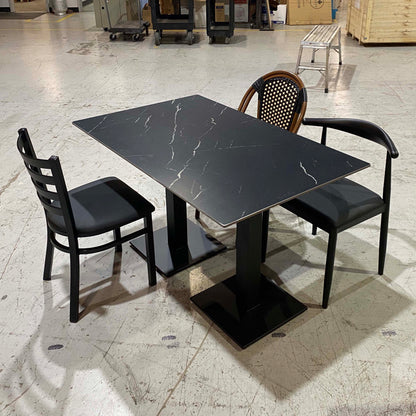 A rectangular black marble-patterned dining table with a black pedestal base, surrounded by three black chairs in a warehouse showroom.