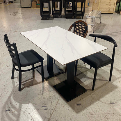 A rectangular dining table with a white marble-patterned top and black pedestal base, surrounded by three black chairs in a warehouse showroom.