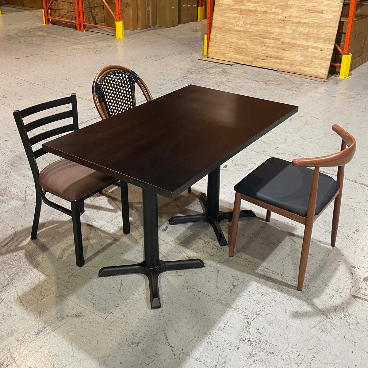 A dark brown rectangular table with a black pedestal base, shown with two chairs in a warehouse showroom setting.