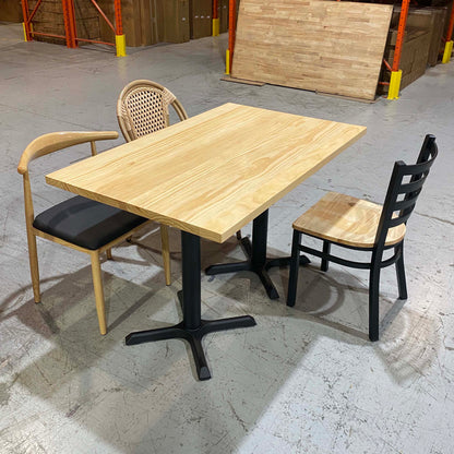 A rectangular light wood dining table with a black metal pedestal base, shown with two chairs in a warehouse-like setting. One chair has a light wood frame and black seat, and the other has a woven back and light wood seat.
