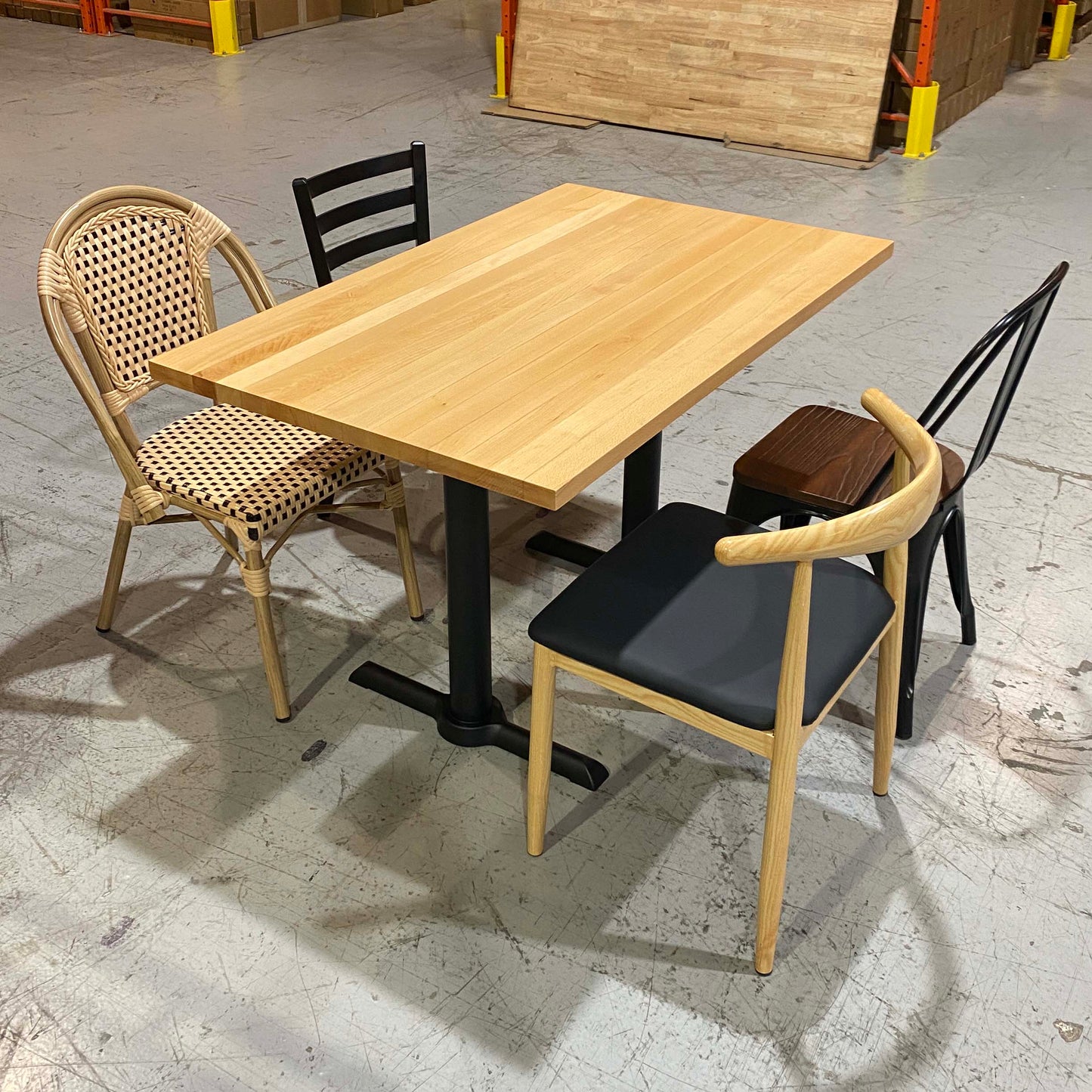 A rectangular light wood dining table with black metal pedestal legs, surrounded by three chairs: a woven rattan chair with a beige seat, a black chair with a dark cushion, and a light wood chair with a black cushion. The scene is photographed in a warehouse showroom with concrete floors and storage items in the background.