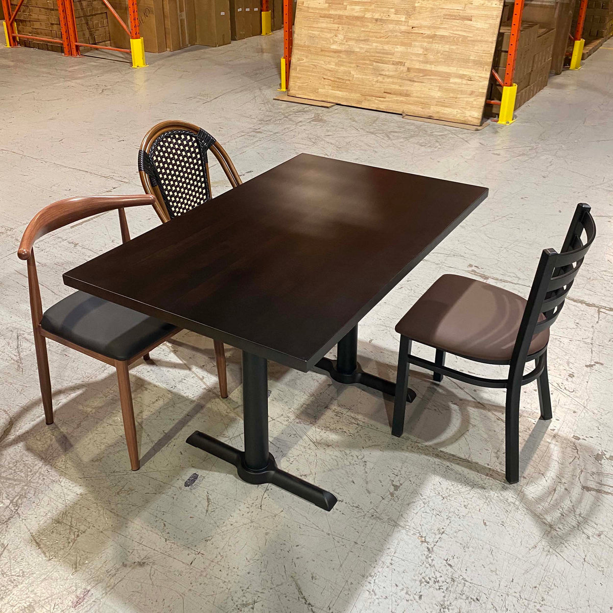 A dark brown rectangular restaurant table with a pedestal base, shown with two chairs in a warehouse showroom setting.