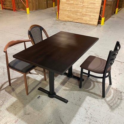 A dark brown rectangular restaurant table with a pedestal base, shown with two chairs in a warehouse showroom setting.