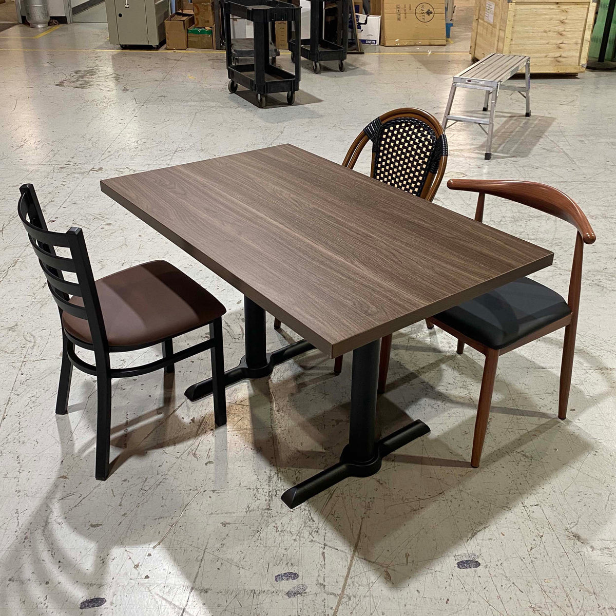 A rectangular wood-look dining table with a dark brown top and black metal pedestal base, shown with three chairs in a warehouse showroom setting.