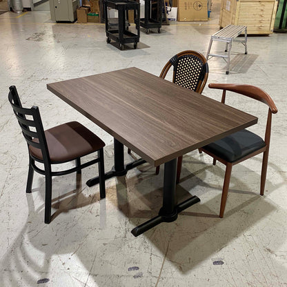 A rectangular wood-look dining table with a dark brown top and black metal pedestal base, shown with three chairs in a warehouse showroom setting.