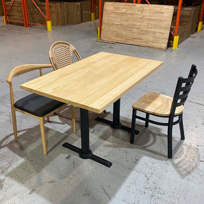 A light natural wood rectangular table with black metal pedestal legs, shown with two chairs in a warehouse setting. One chair has a woven back and black seat cushion, and the other has a black slatted back and light wood seat.