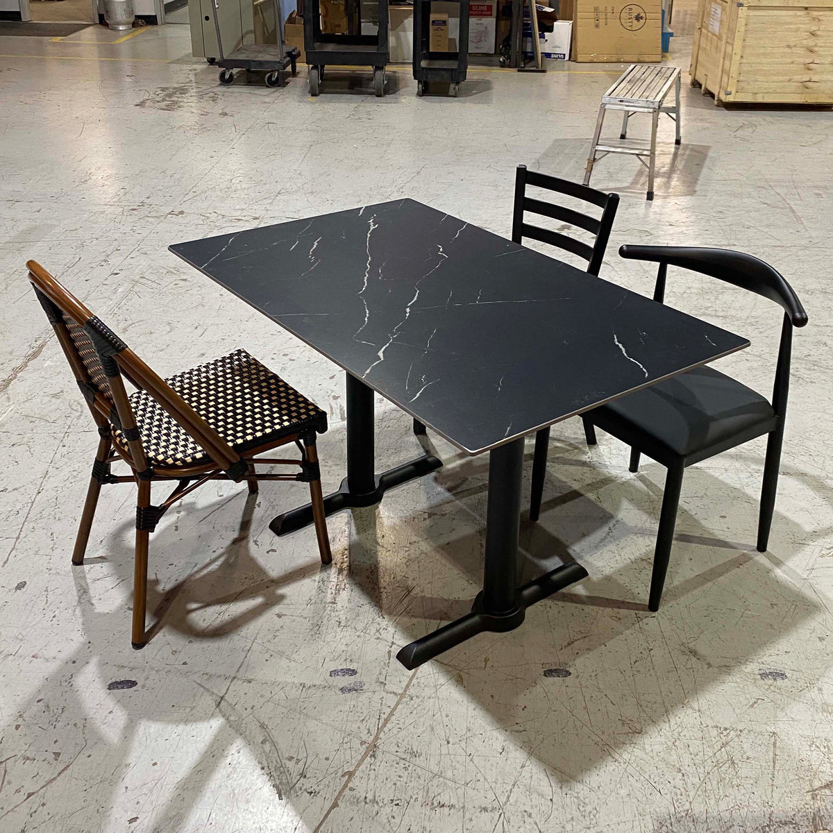 A rectangular black dining table with a white marble-look veining pattern, accompanied by two chairs in a warehouse setting. One chair is black with a curved backrest, and the other is a brown wooden folding chair with a woven seat.