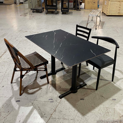 A rectangular black dining table with a white marble-look veining pattern, accompanied by two chairs in a warehouse setting. One chair is black with a curved backrest, and the other is a brown wooden folding chair with a woven seat.