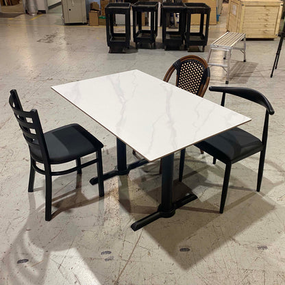 A square white marble-patterned dining table with a black pedestal base, surrounded by three black chairs in a warehouse showroom.