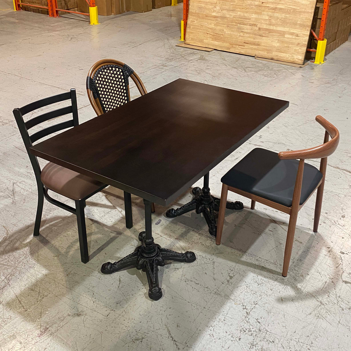 A dark brown rectangular dining table with an ornate black metal pedestal base, accompanied by two chairs in a warehouse showroom setting.
