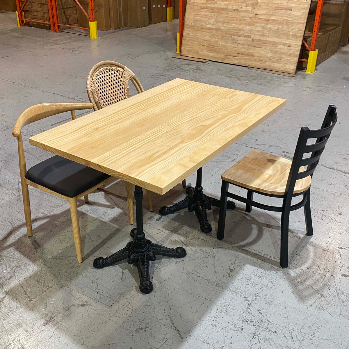 A rectangular light wood dining table with black pedestal legs, shown with two chairs in a warehouse-like setting with concrete floors and wooden panels in the background.
