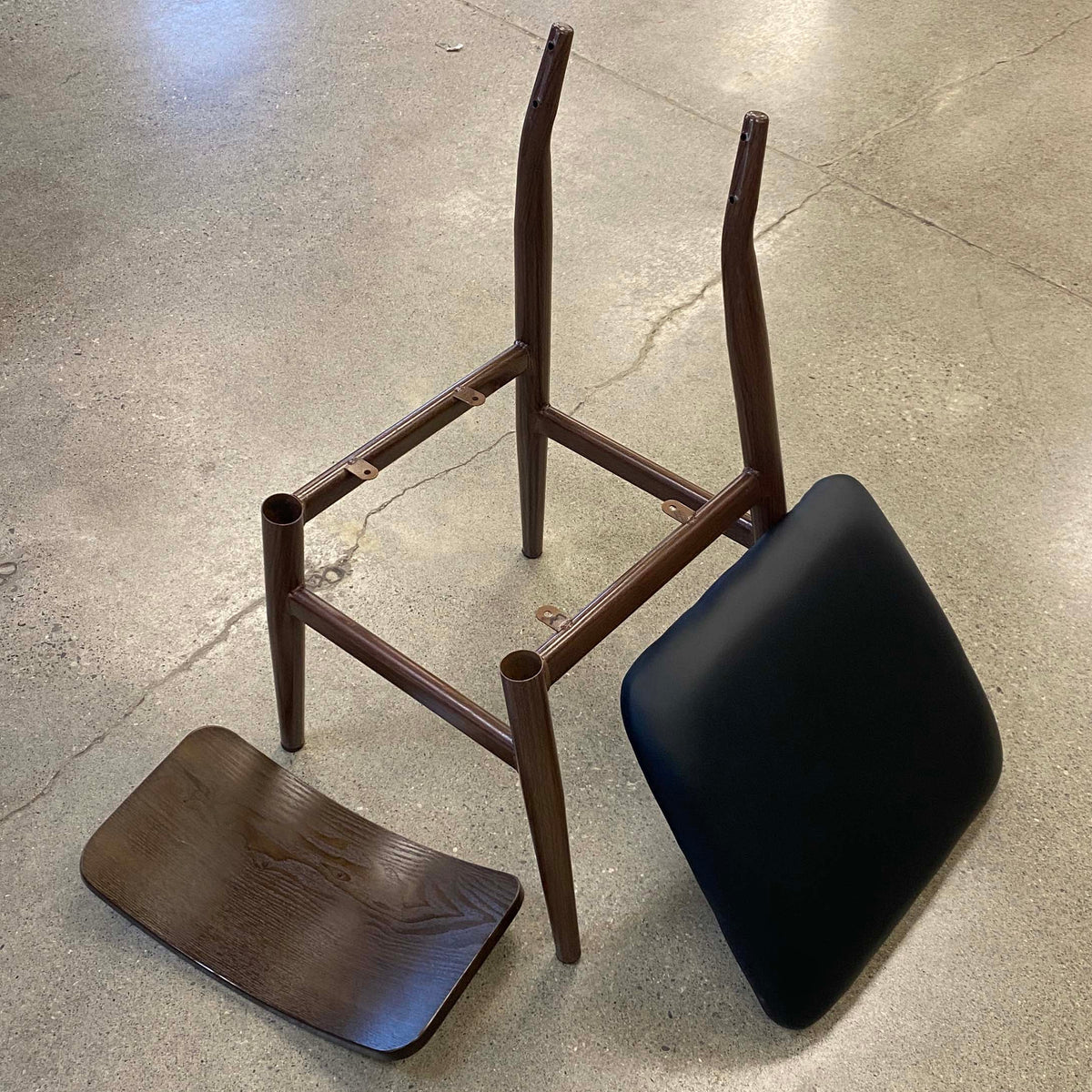 A metal chair frame with a dark brown finish, shown disassembled with its seat and backrest placed beside it on a concrete floor.