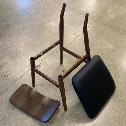A metal chair frame with a dark brown finish, shown disassembled with its seat and backrest placed beside it on a concrete floor.
